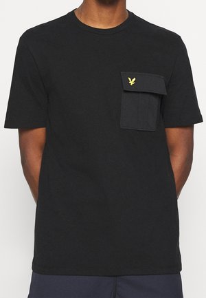 Sort t-shirt med korte ærmer og en flap-lomme på venstre bryst med en lille gul fuglelogo, båret af en person med armene bag ryggen.