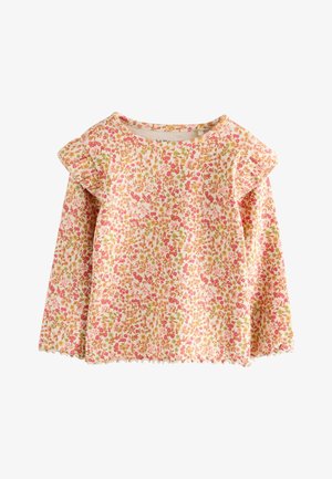 Top de flores de manga larga con volantes en los hombros, combinado con pantalones acampanados rosa con textura acanalada y zapatillas blancas. Fondo minimalista con formas suaves.