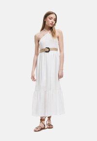 Robe maxi blanche sans manches avec un corsage smocké et un ourlet en volants. Elle est dotée d'une ceinture tissée avec une boucle ronde. Le modèle porte des sandales marron.