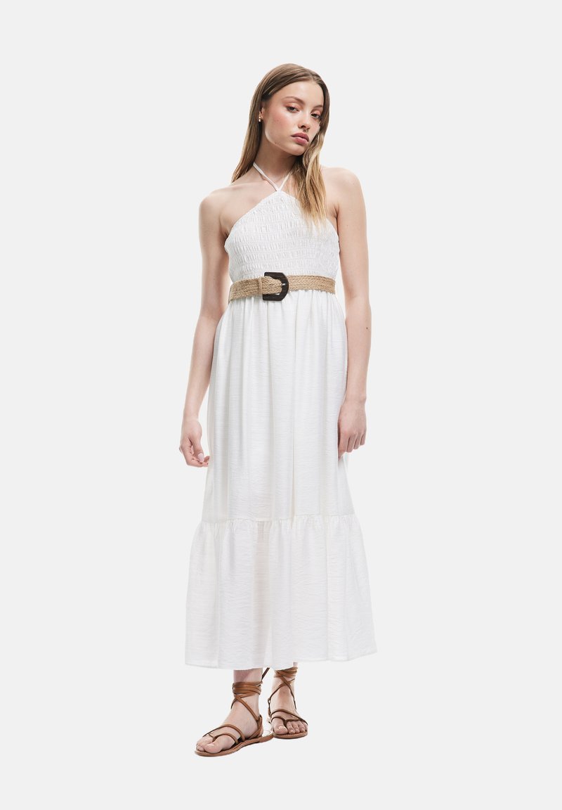 Robe maxi blanche sans manches avec un corsage smocké et un ourlet en volants. Elle est dotée d'une ceinture tissée avec une boucle ronde. Le modèle porte des sandales marron.