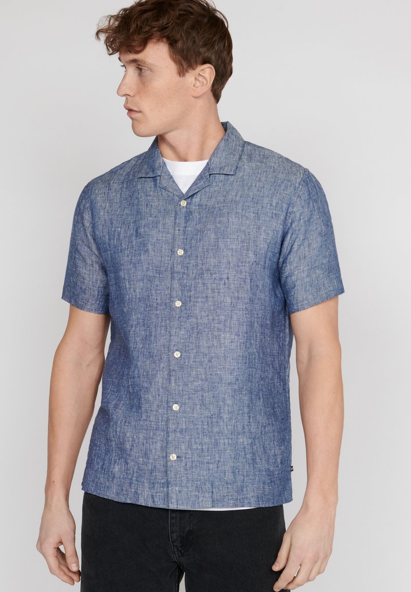 Camicia a maniche corte con bottoni in un tessuto gradient blu texture. Presenta un colletto da campeggio e bottoni di colore chiaro per contrasto.
