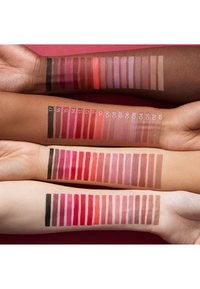 Quatre bras avec des teints de peau variés présentent chacun 17 échantillons de rouge à lèvres numérotés, allant du noir et des rouges profonds aux roses clairs et aux marrons.