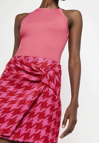 Růžový žebrovaný halenka bodysuit spárovaný s červeno-růžovou sukní s houndstooth vzorem, která má uzlový detail v pase.