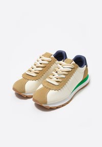 Zapatillas beiges y blancas con un acento verde, que presentan un diseño con borde en forma de concha, ante texturizado y cordones blancos sobre una suela de goma.