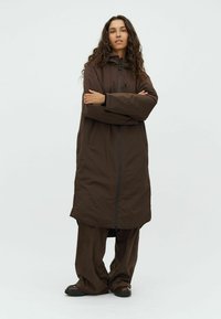 Bruine lange jas met capuchon, frontale rits en zijzakken; gecombineerd met ruime bruine broek en zwarte schoenen. Gladde textuur.