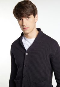 Cardigan a coste scuro con colletto a scialle, dotato di chiusura con bottoni e due tasche frontali, indossato sopra una camicia bianca.