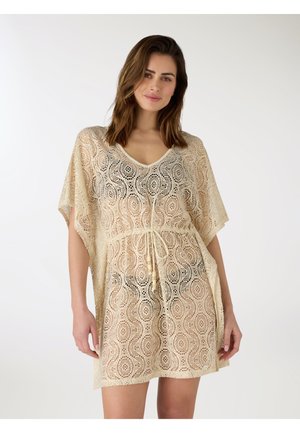 Kobieta w beżowej szydełkowanej narzutce plażowej z dekoltem w serek i sznurkiem w talii, stojąca na jednolitym białym tle.