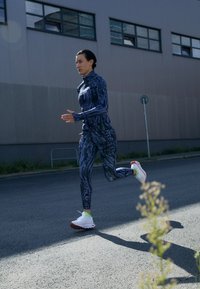 Tamsiai mėlyna sportinė apranga su abstrakčiomis raštais, prigludęs ilgomis rankovėmis marškinėliai ir legginsai, derinami su baltais ir raudonais bėgimo batais.