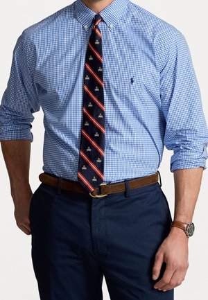 Mann trägt ein blau kariertes Hemd mit hochgekrempelten Ärmeln, eine marineblaue gestreifte Krawatte, einen braunen Gürtel, dunkle Hose und eine Armbanduhr am linken Handgelenk.