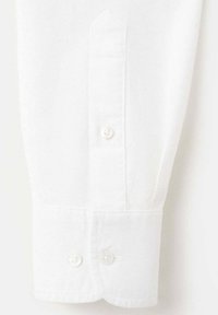 Poignet de chemise en coton blanc avec une extrémité pointue, équipé de deux boutons ronds blancs et un détail de couture visible. Texture douce et couleur uniforme.