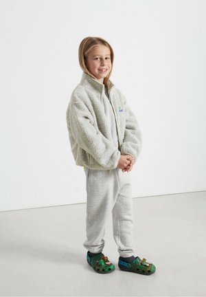 Grijze fleece zipjack, grijze sweatshirt, grijze joggingbroek en groene schoenen met clip-on accessoires. Soepele textuur; casual, ontspannen pasvorm.