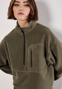 Olivengrøn fleece-pullover med høj krave, halv lynlås foran og lynlåslomme på venstre bryst. Blød tekstur og afslappet pasform.