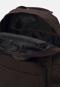 Mochila marrón oscuro con una textura de tela suave, que cuenta con una abertura con cremallera y un interior dividido con un forro negro y un bolsillo.