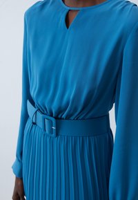 Blå veckad klänning med långa ärmar, som har en smal nyckelhålsringning och ett matchande bälte. Tillverkad av slät tyg med texturerade accenter.