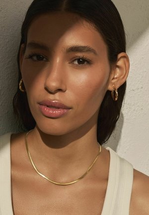 Woman with medium skin tone and dark hair wears gold hoop earrings and a thin gold necklace, lit by natural light against a textured wall.