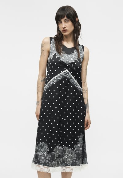 Γυναίκα που φοράει αμάνικο μαύρο φόρεμα με λευκές πουά, δαντελένια λουλουδάτα σχέδια και λεπτομέρεια φιόγκου, στέκεται μπροστά σε απλό φόντο.
