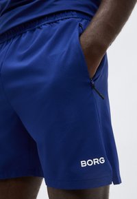 Κοντινή λήψη του χεριού ενός ατόμου στην τσέπη μπλε αθλητικών σορτς με φερμουάρ και λογότυπο «BORG» στο πόδι.
