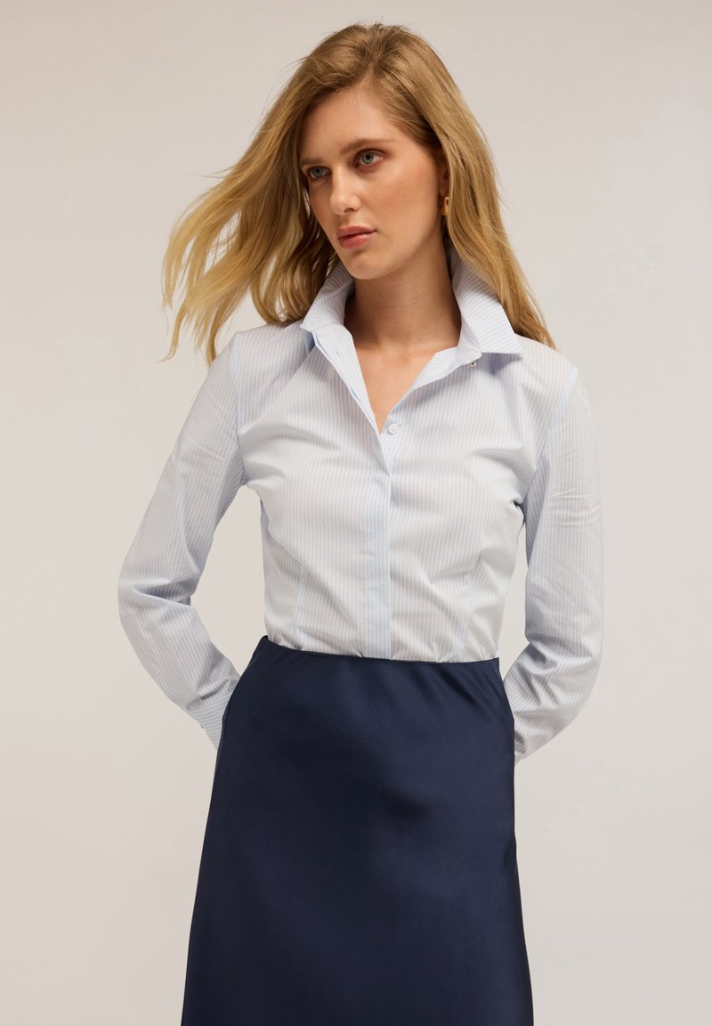 Camicia button-up a righe blu chiaro con colletto aperto, abbinata a una gonna a tinta unita di un blu navy scuro. Il tessuto è liscio e leggermente aderente.