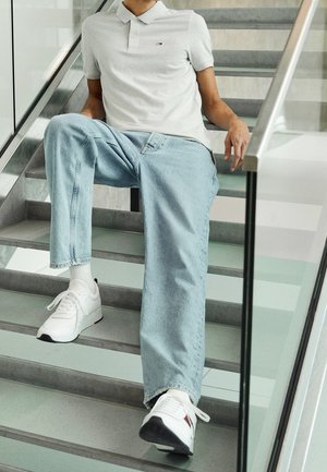 Person sidder på indendørs metaltrappe iført lyseblå jeans, hvid poloshirt, hvide sneakers og hvide sokker.