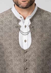 Grau gemusterte Weste mit Silberknöpfen und Tasche, über einem weißen Hemd mit Kragen, das eine Kettenakzentuierung aufweist. Strukturierte Stoffdetails sind sichtbar.