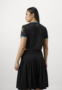 Jersey preto cropped com mangas curtas, com detalhes em azul e amarelo, combinado com uma saia preta plissada. Visão traseira em destaque.
