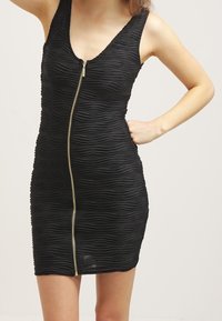 Černé bezramínkové šaty s texturovaným vlnitým vzorem, s předním zlatým zipem. Přiléhavý střih, délka ke kolenům, hladká tkanina.