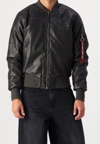 Černá kožená bomber bunda s ribkovanými manžetami a lemem. Má zapínání na zip a kapsu na rukávu s červeným štítkem.