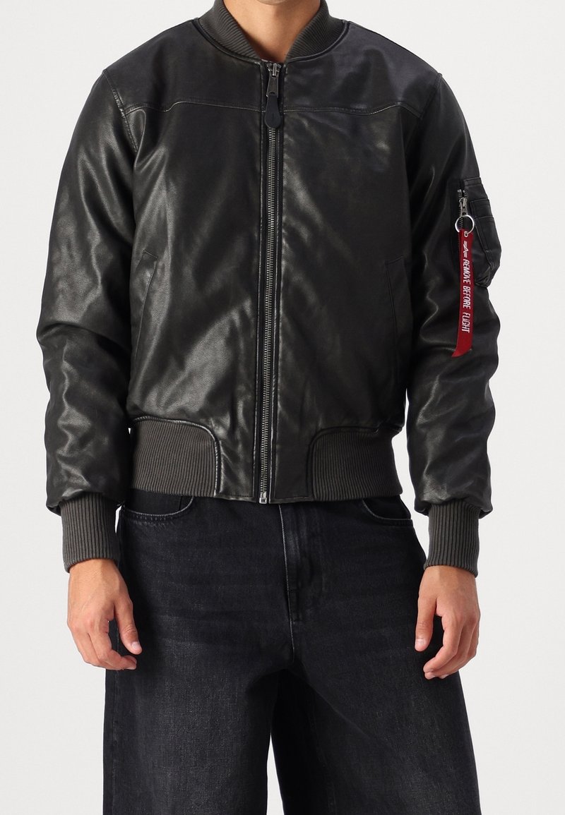 Černá kožená bomber bunda s ribkovanými manžetami a lemem. Má zapínání na zip a kapsu na rukávu s červeným štítkem.
