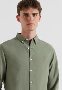 Groene overhemden gemaakt van lichte stof met een kraag, voorzien van witte knopen en een geborduurd gouden logo op de borst.