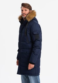 Cappotto imbottito blu navy con cappuccio bordato di pelliccia, tasche frontali e accentature nere sulle spalle. Presenta una chiusura con zip e bottoni.