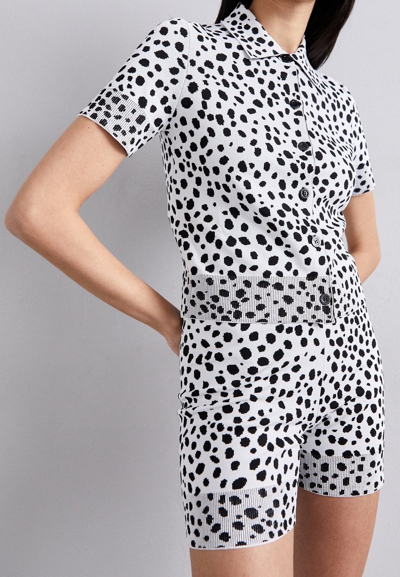 Femme portant un haut blanc à manches courtes boutonné et un short assorti avec un motif à pois noirs irréguliers, debout les mains sur les hanches.