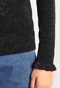 Svart stickad tröja med metalltråd; har rufflade ärmslut och en åtsittande design. Kombinerad med blå denim.