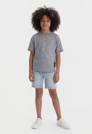 Enfant aux cheveux bouclés debout portant un t-shirt gris à manches courtes, un short en jean clair et des baskets blanches sur un fond uni.