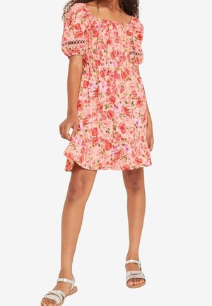 Niña con un vestido rosa floral sin hombros con mangas abullonadas y sandalias blancas, posando frente a un fondo liso.