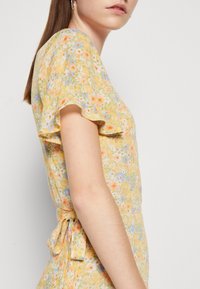 Robe fleurie jaune avec des manches courtes à volants, dotée d'un lien à la taille et d'un tissu texturé avec des motifs multi-couleurs complexes.