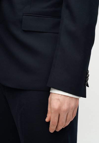 Χέρι ακουμπισμένο στο πλάι, ντυμένο με σκούρο επίσημο κοστούμι με ορατό λευκό μανικετόκουμπο που δείχνει τα κουμπιά του μανικιού και μια πλαϊνή τσέπη.