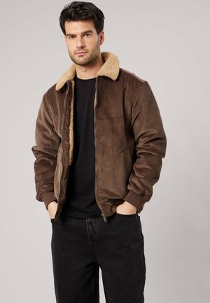 Man met donker haar die een bruin ribfluwelen jas draagt met een beige schapenvachtkraag en een zwart overhemd en broek, staand met handen in de zakken.