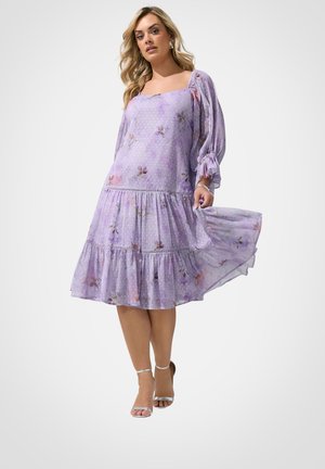 Vrouw draagt een lichtpaarse bloemen midi-jurk met pofmouwen en een vierkante halslijn, houdt de rok van de jurk vast terwijl ze staat.