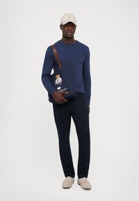Jūrų laivyno ilgomis rankovėmis marškiniai, tamsios kelnės, smėlio spalvos šlepetės ir smėlio spalvos kepurė. Laikosi tamsaus rankinuko, ant kurio pavaizduotas lokys jūreivio aprangoje.
