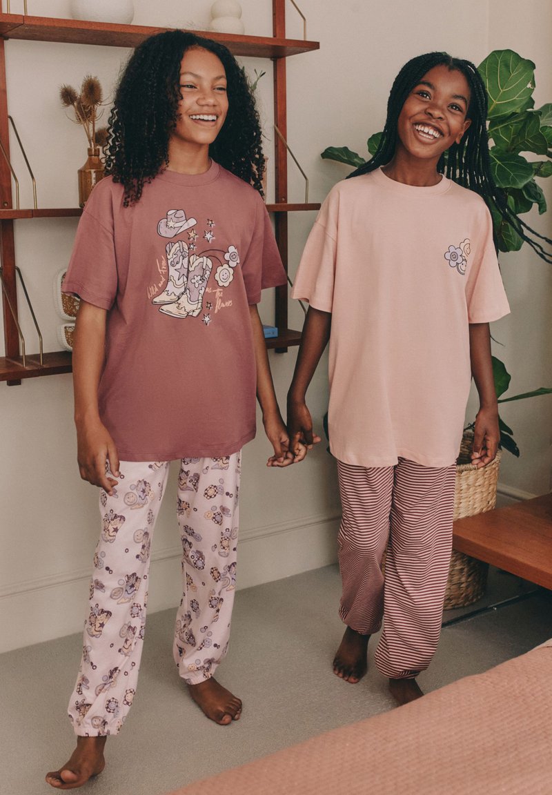 Deux enfants se tiennent la main, portant des t-shirts graphiques et des pantalons de pyjama à motifs. Un t-shirt est bordeaux avec un dessin, l'autre est rose pâle.