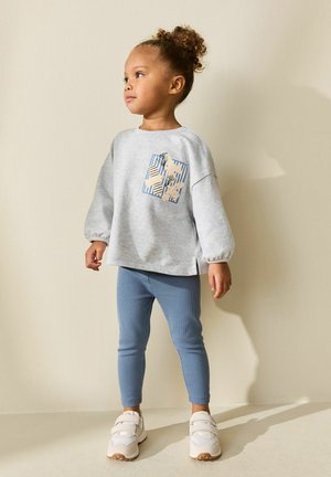 Ungt barn med krøllet hår står iført en lysegrå sweatshirt med blomsterprint, blå leggings og hvide sneakers mod en beige væg.