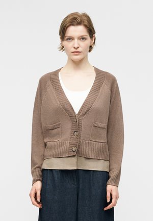 Ung kvinde med kort hår iført en brun knap-foran cardigan over en hvid top og mørke bukser, stående mod en ensfarvet baggrund.
