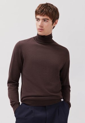 Ung mand iført en brun rullekrave og mørkeblå bukser, står med hænderne i lommerne og kigger sidelæns.