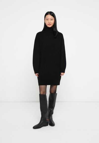 Femme se tenant dans une robe pull côtelée noire avec des collants transparents et des bottes en cuir noires montantes avec des détails dorés.