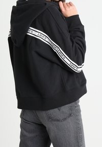 Sudadera negra recortada con capucha ajustable. Presenta rayas de marca blanca a lo largo de las mangas. El material parece suave y tiene un corte relajado.