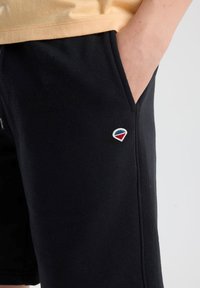 Black shorts made of soft fabric feature side pockets and a small embroidered logo in red, blue, and white on the lower left side.
