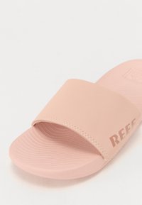 Rosa sklisandal laget av glatt materiale, med en bred rem og rillede såler; har merkevarenavnet "REEF" embossert på siden.