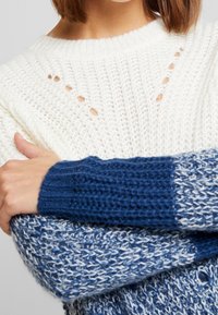 Stickad tröja med vit kropp, mörkblå ribbade muddar och ljusblå detaljer. Designen inkluderar öppna stickade detaljer vid halsringningen.
