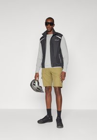 Schwarze ärmellose Weste über einem grauen Langarmshirt, khakifarbene Shorts, schwarze Schuhe, hält einen grauen Helm, mit dunklen Sonnenbrillen und sichtbarem Branding.