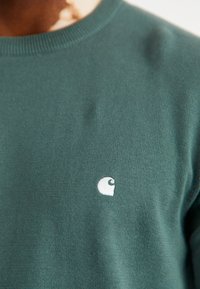 Pull en maille verte avec un col rond. Présente un petit logo blanc sur la poitrine. Le matériau affiche une finition lisse et texturée.